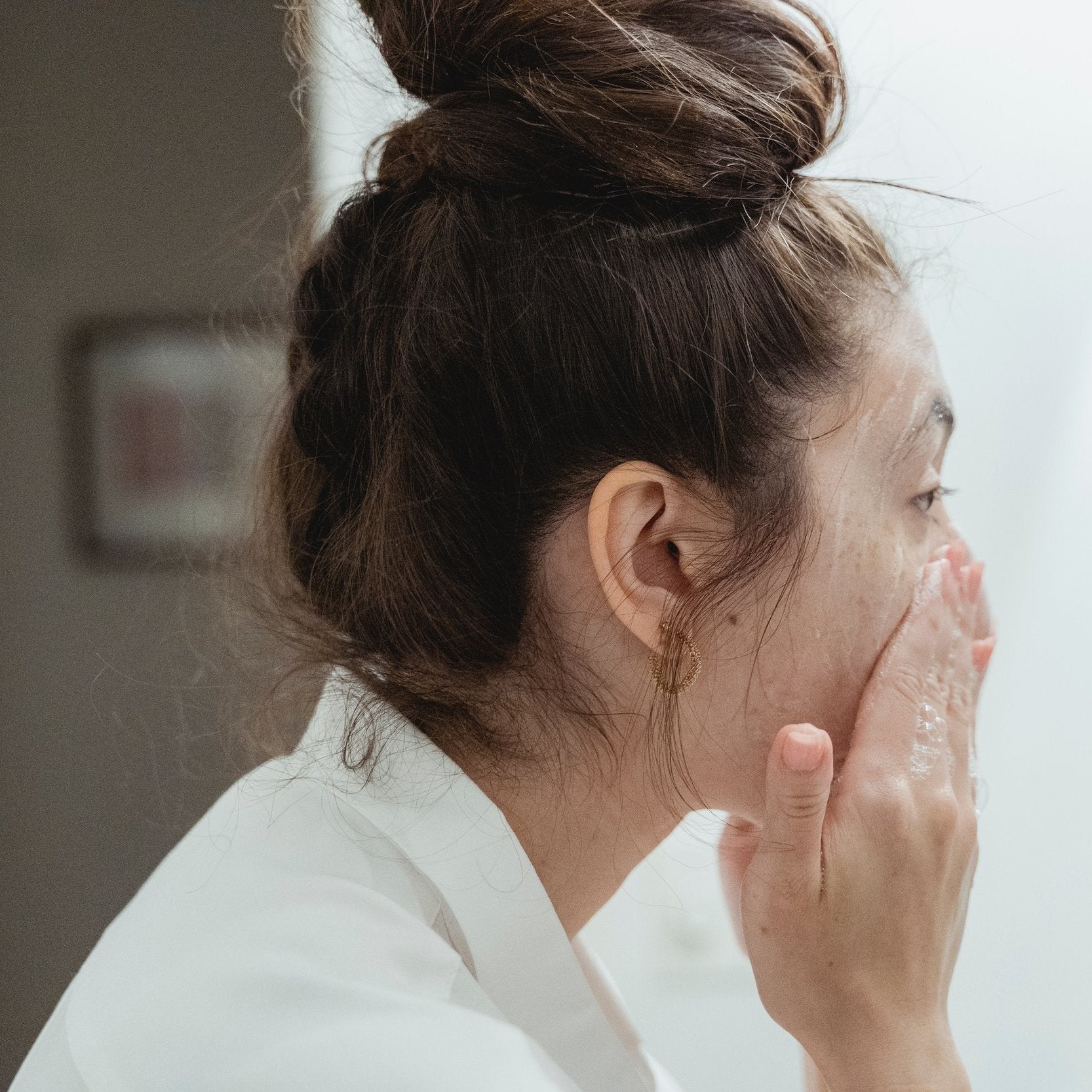Profile of woman with hair in bun cleansing her face.