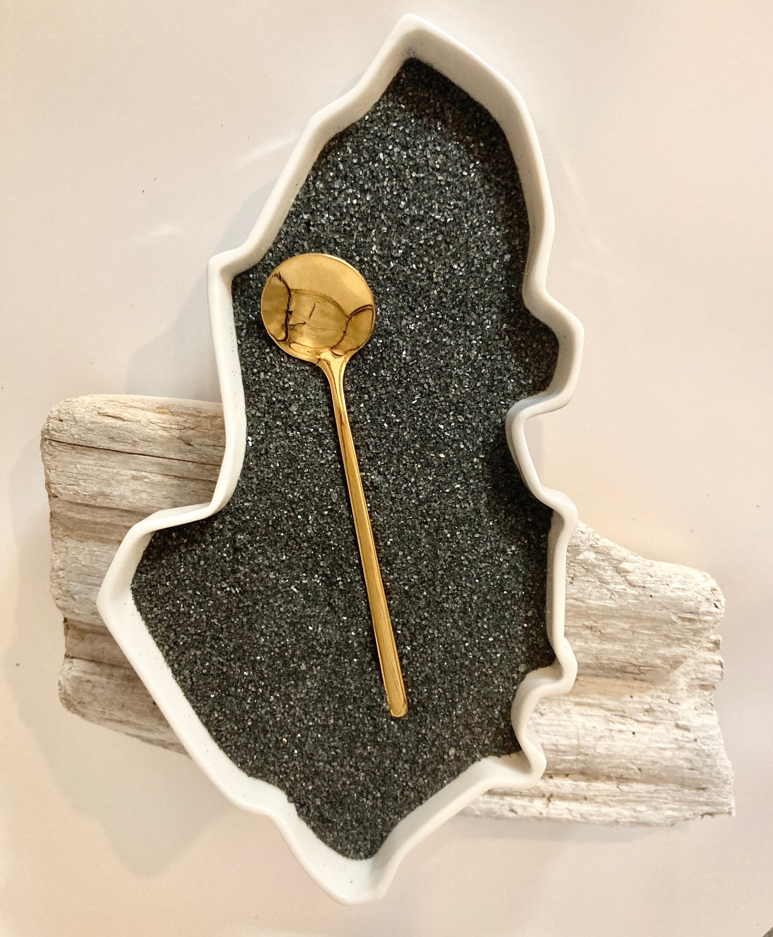 Gold spoon on ground black charcoal in an amorphous white bowl.