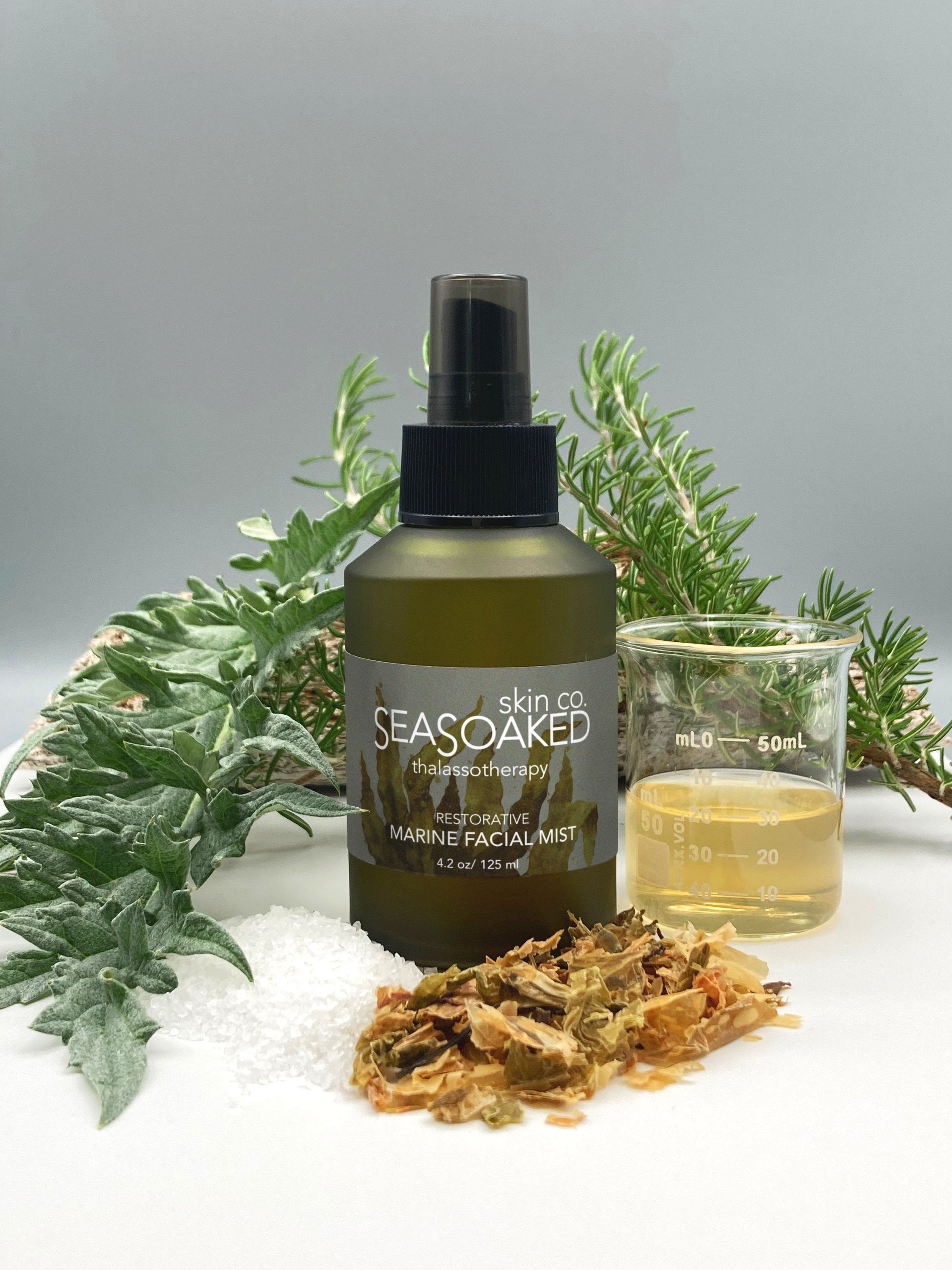 Marine Facial Mist spray bottle near beaker of liquid, seaweed, and other natural ingredients.